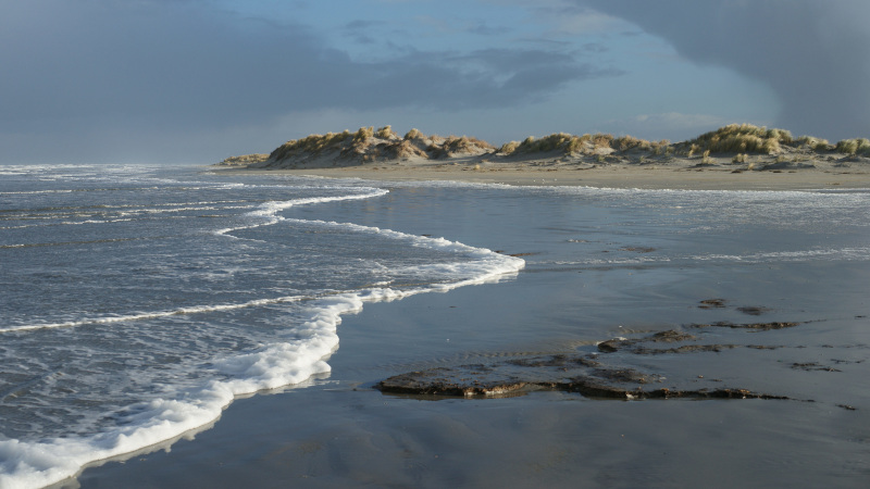 De zee komt tot aan de duinen en overspoeld het strand. Op de voorgrond een stuk klei in het zand.
