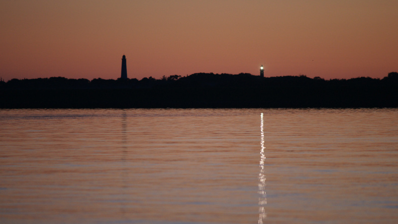 Silhouetten van twee torens met zee op de voorgrond, gehuld in oranje avondlicht en de lamp van de vuurtoren weerspiegeld in het water.