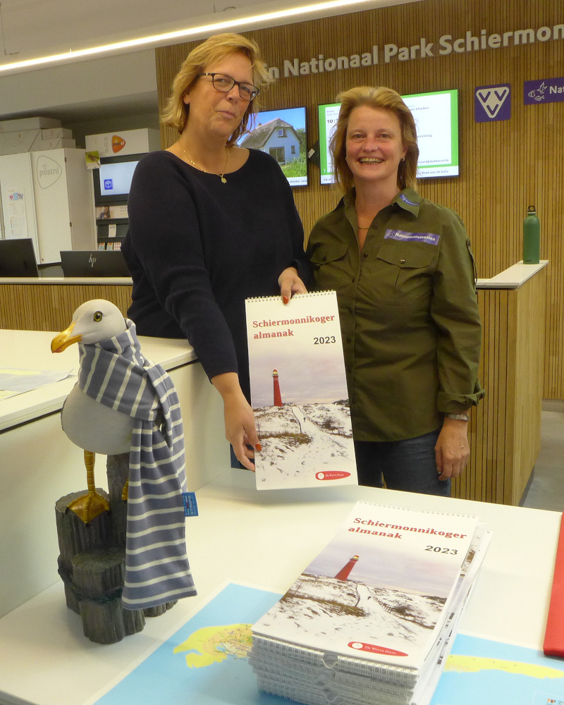 Twee dames presenteren de kalender met opschrift Schiermonnikoger almanak 2023 aan de balie van het informatiecentrum