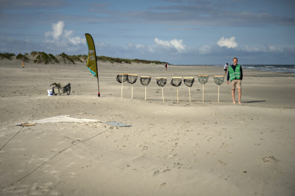 Man in groen hesje op het strand naast acht schepnetjes, een groene-strand-beach flag en een fietskar.