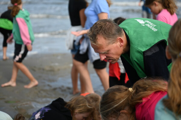 Een volwassene in groen hesje buigt zich over kinderen die gebogen staan over iets wat op het strand ligt.