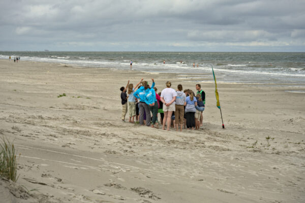 groep kinderen op het strand met begeleiders. Vier kinderen steken een vinger omhoog.