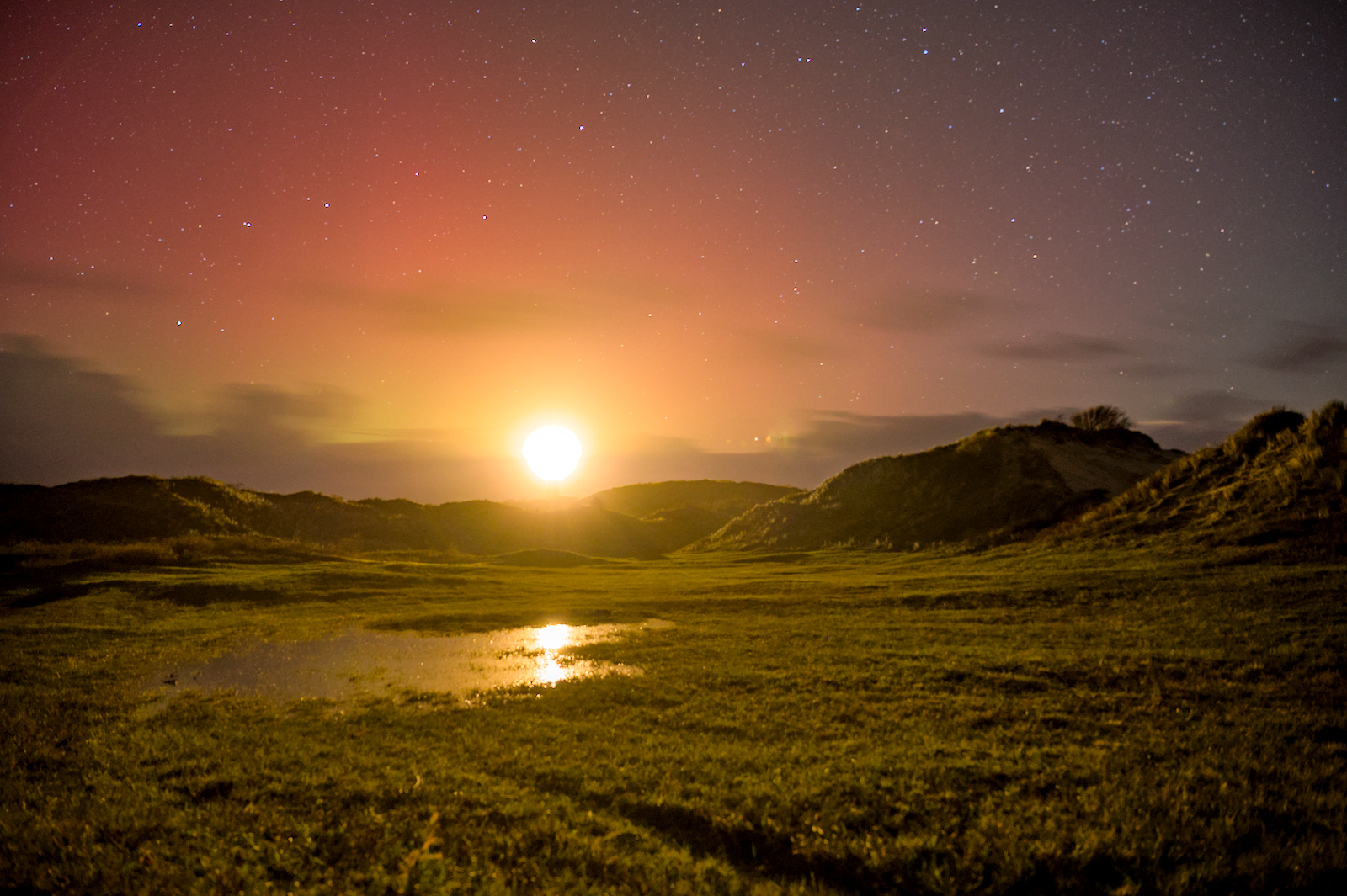 Fel licht boven een duinenrij weerkaatst in een plas in de duinvallei. Een rood-oranje gloed aan de nachtelijke sterrenhemel.