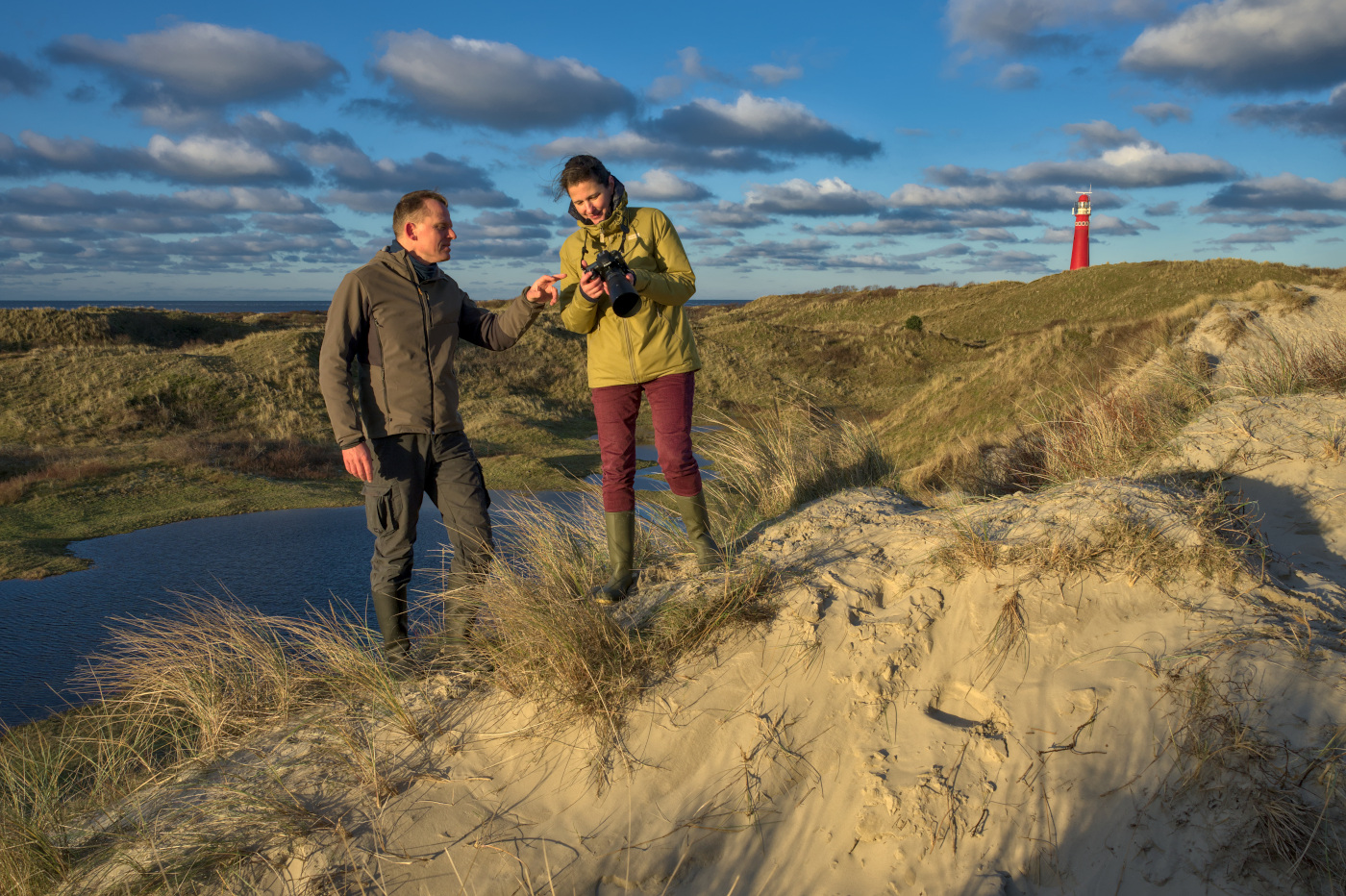 een man en een vrouw staan op een duin. De vrouw heeft een camera in de handen, de man wijst ernaar. Op de achtergrond zee en rode vuurtoren.
