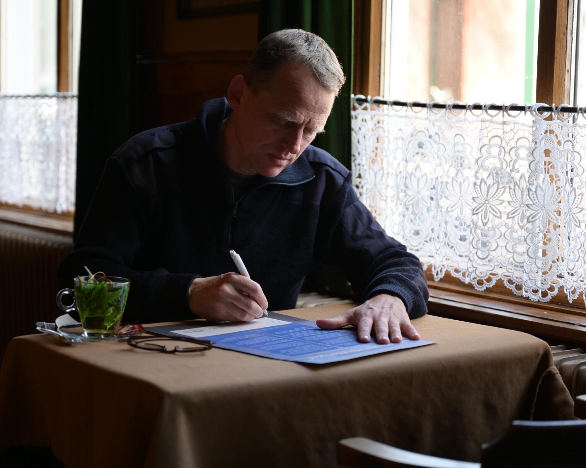 Een man tekent een oorkonde aan een tafeltje met bruin tafelkleed bij het raam.
