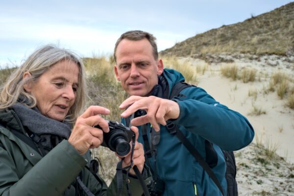 Een man en vrouw wijzen naar een camera in een duinenlandschap.