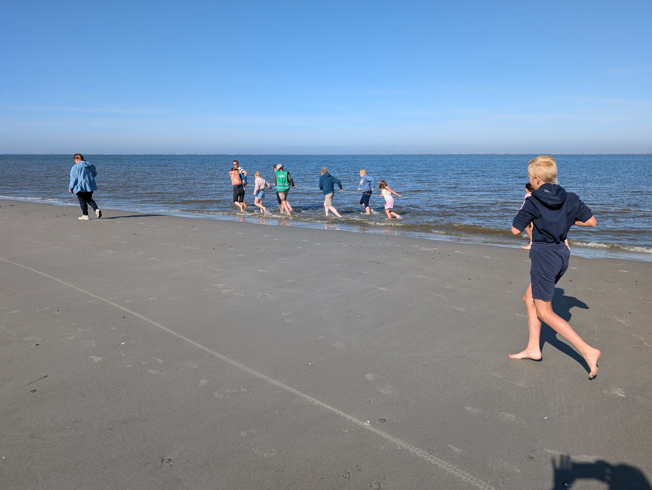 kinderen en volwassenen trekken aan een lang touw en lopen blootvoets door de branding.