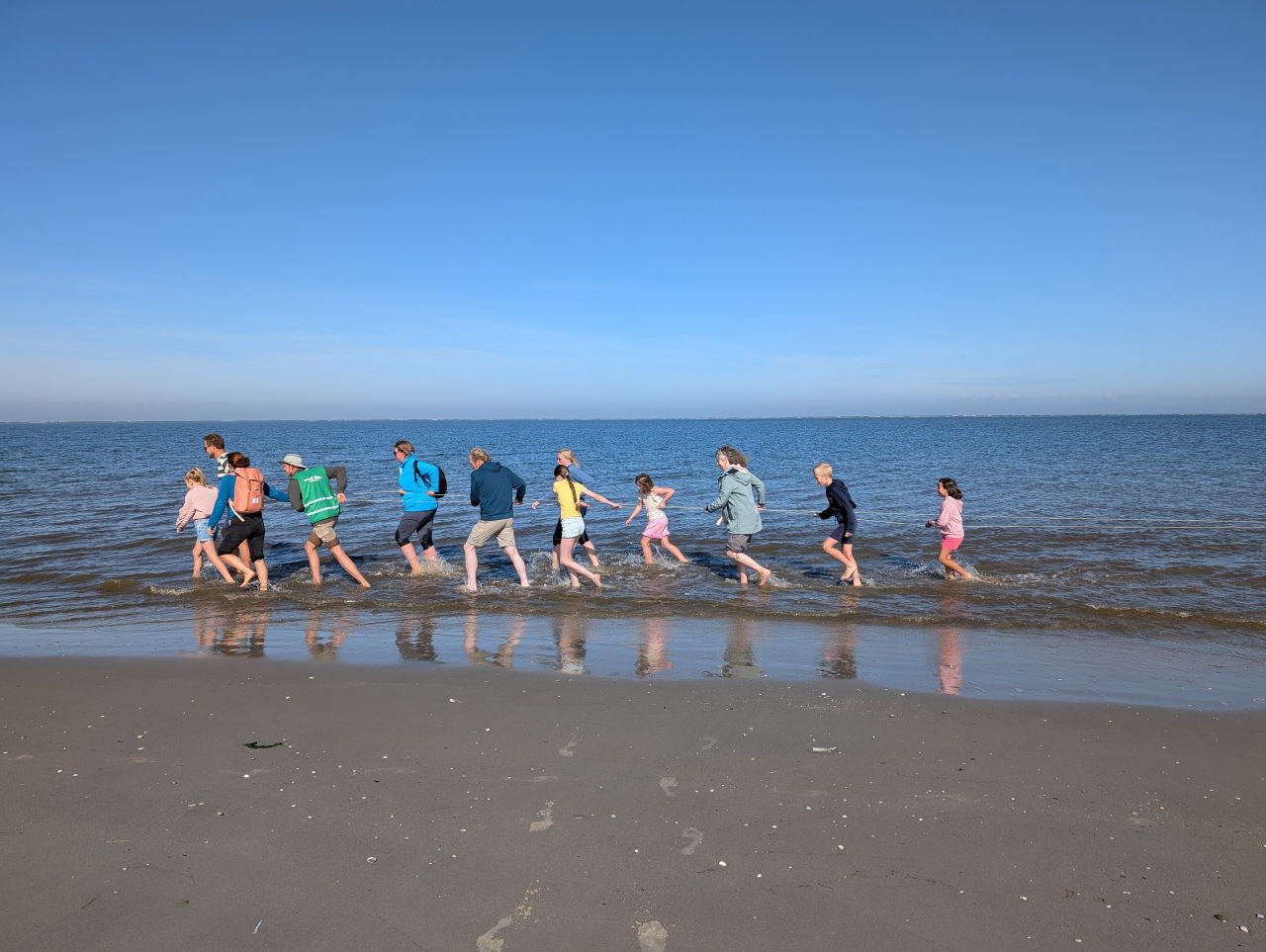 kinderen en volwassenen trekken aan een lang touw en lopen blootvoets door de branding.