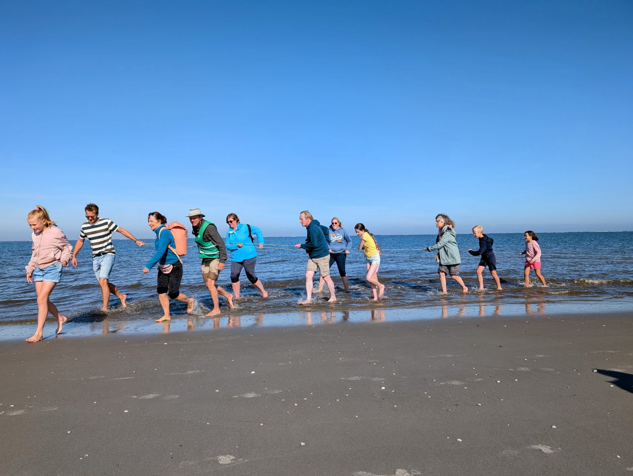 kinderen en volwassenen trekken aan een lang touw en lopen blootvoets door de branding.