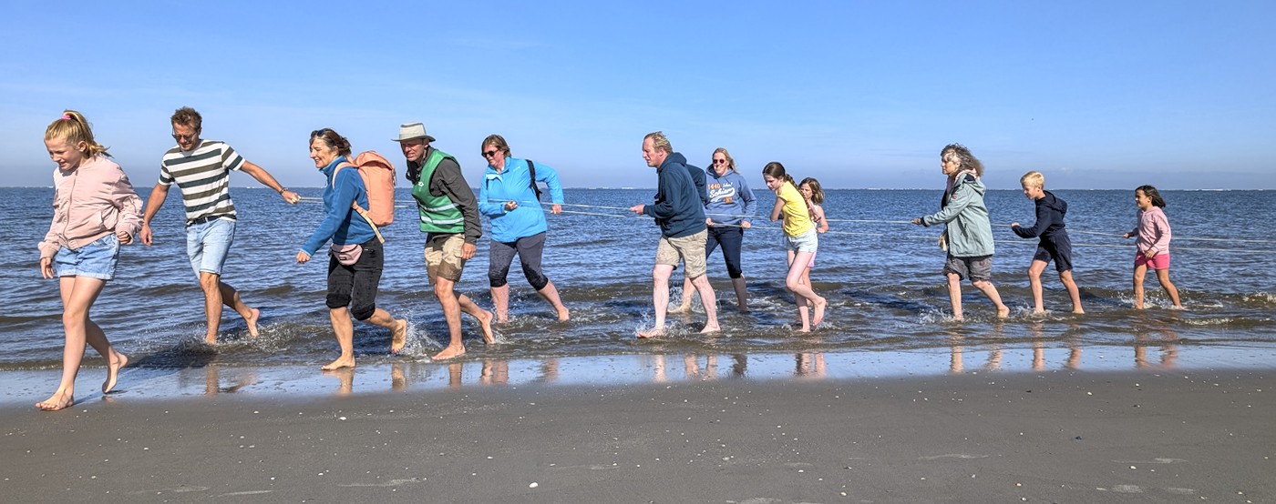 kinderen en volwassenen trekken aan een lang touw en lopen blootvoets door de branding.