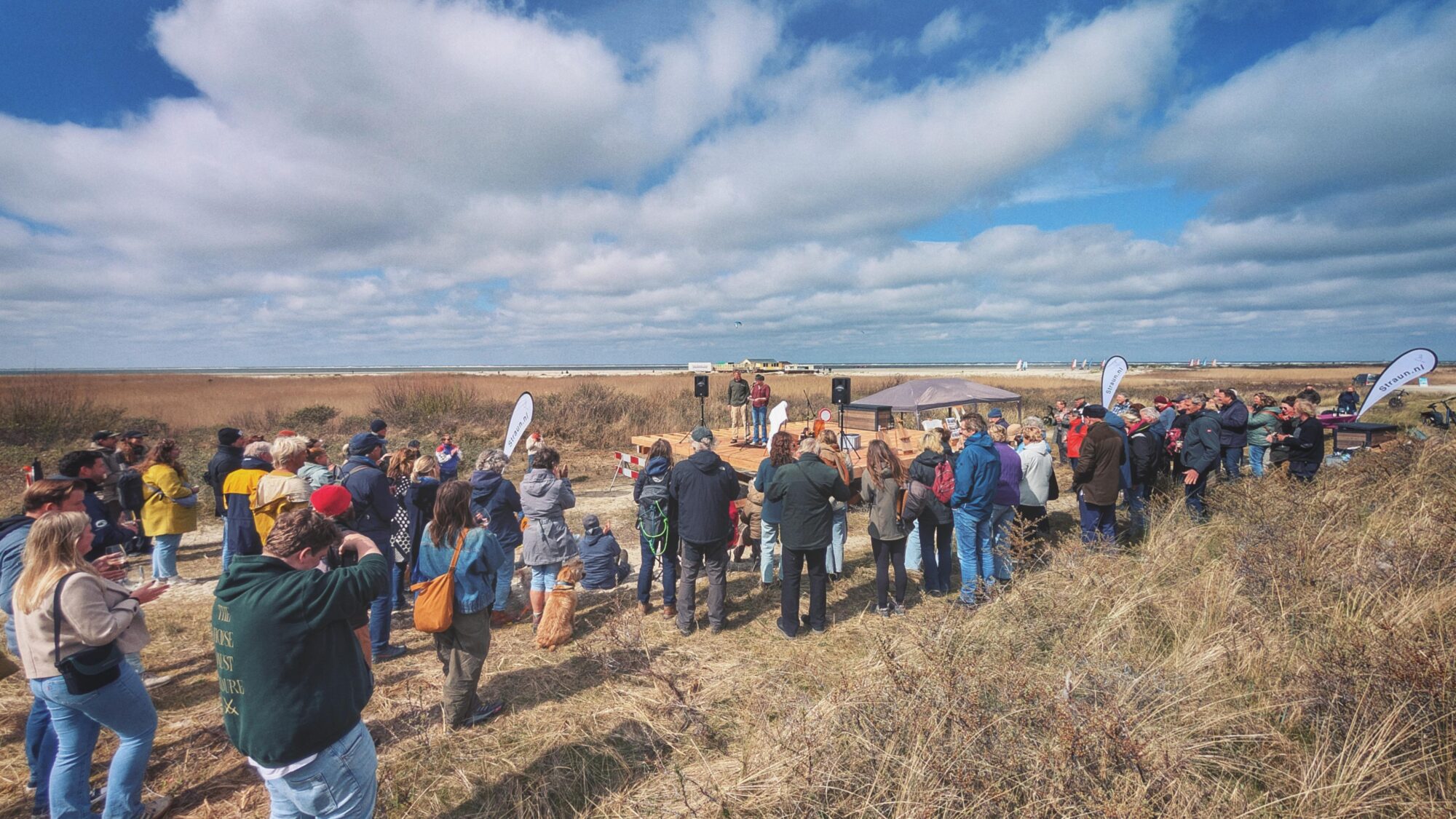 Veel toeschouwers rond een podium op het strand, zonovergoten.